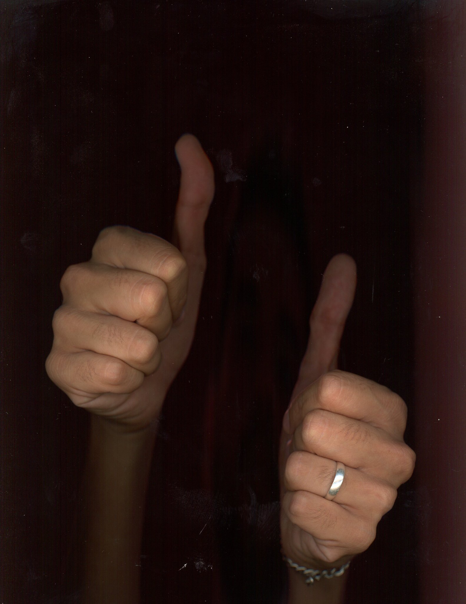 A person showing two fingers, one pointing up and the other towards the side, wearing a ring and a beaded bracelet, on a dark background.