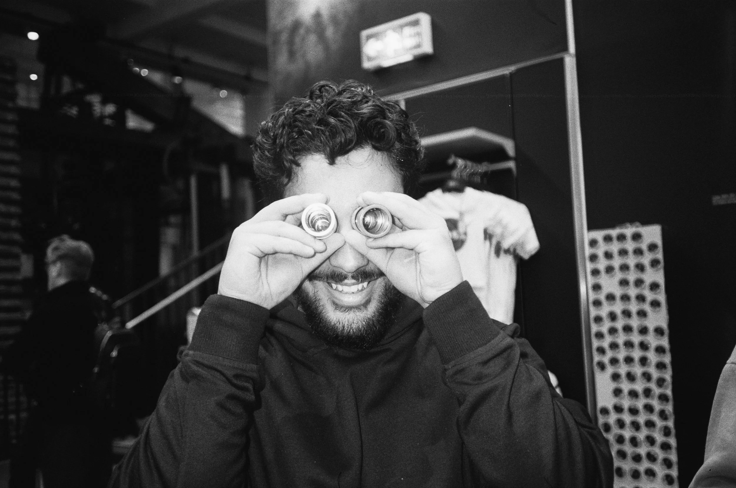 A man with curly hair and a beard looking through binoculars while smiling in a room with dark walls and shelves.
