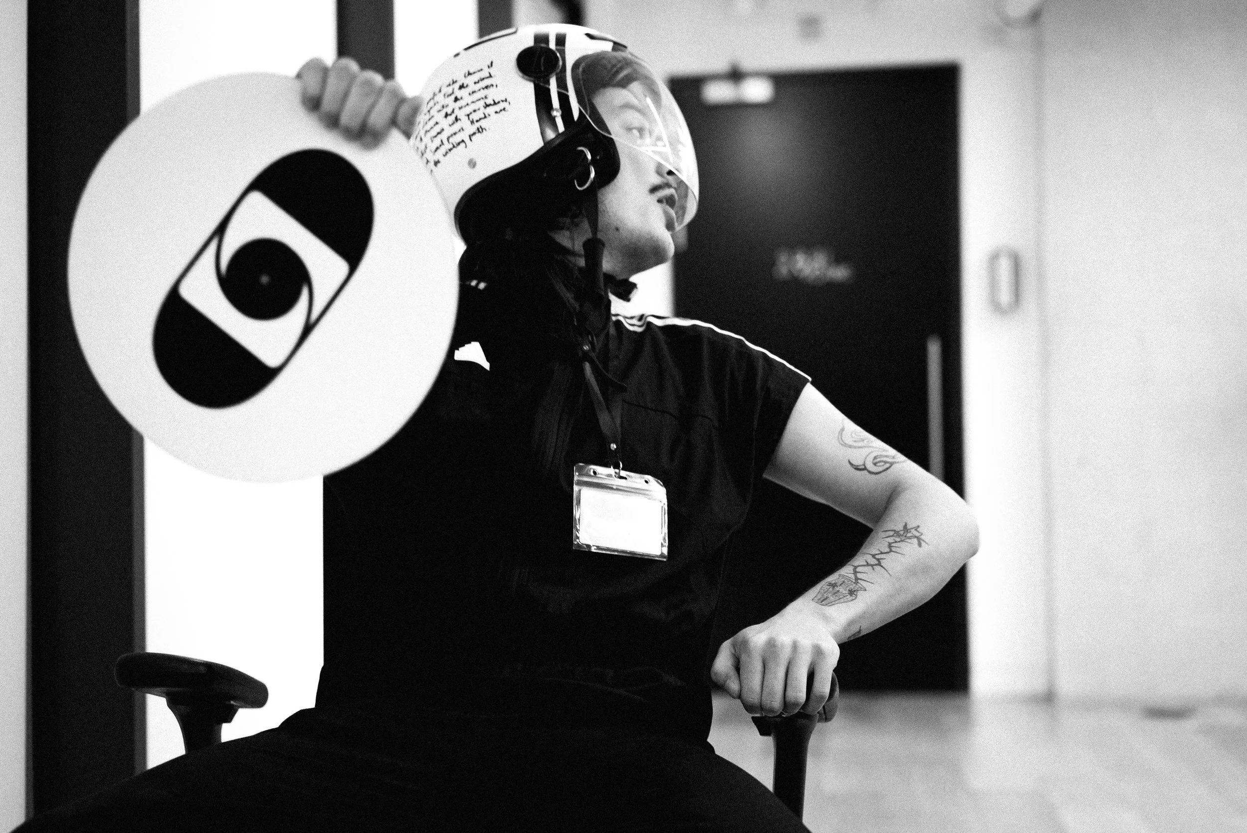 A person sitting in an office chair wearing a helmet with a visor, holding a circular sign with a logo, and wearing a badge around their neck. The person has tattoos on their arm and is dressed in dark clothing. The background is an indoor setting wi