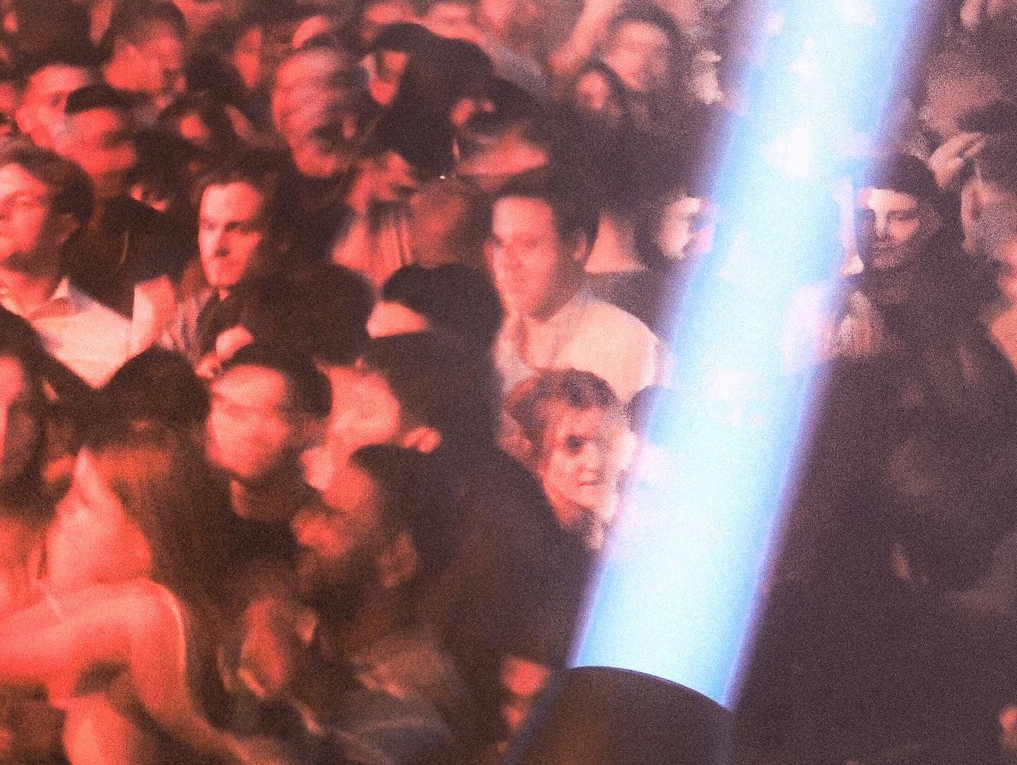Crowd of people attending an indoor event, some wearing glasses and some with noticeably styled hair, with a bright light streak across the image.