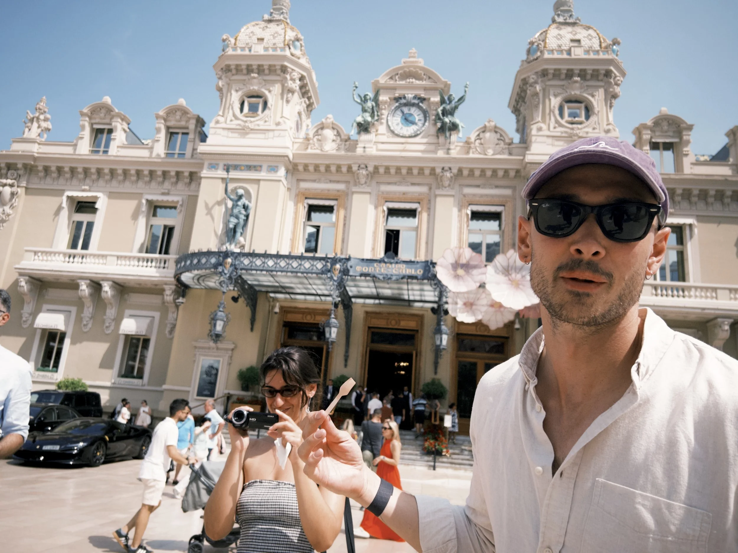 A man in a white shirt and sunglasses taking a selfie in front of a grand building with a clock and sculptures, with a woman nearby holding a camera. There are many people and parked cars around.