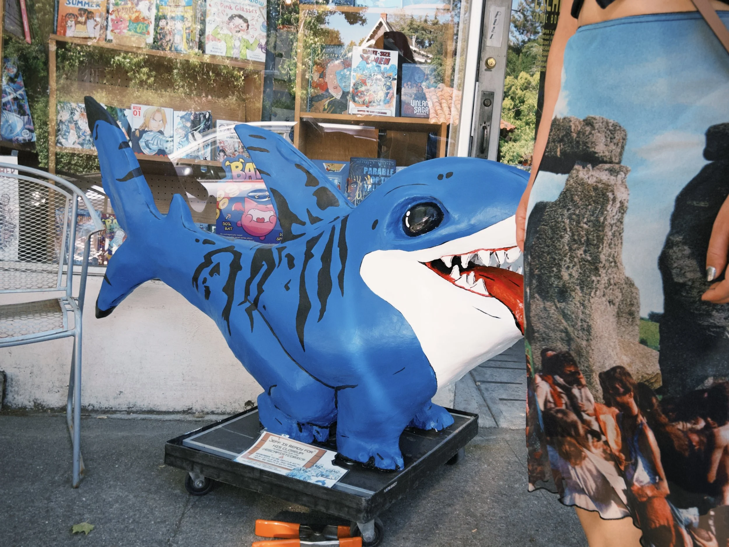 A large blue shark sculpture with open mouth and sharp teeth on display outside a store window featuring comic books and graphic novels. A person wearing a shirt with a scenic print is partially visible on the right.