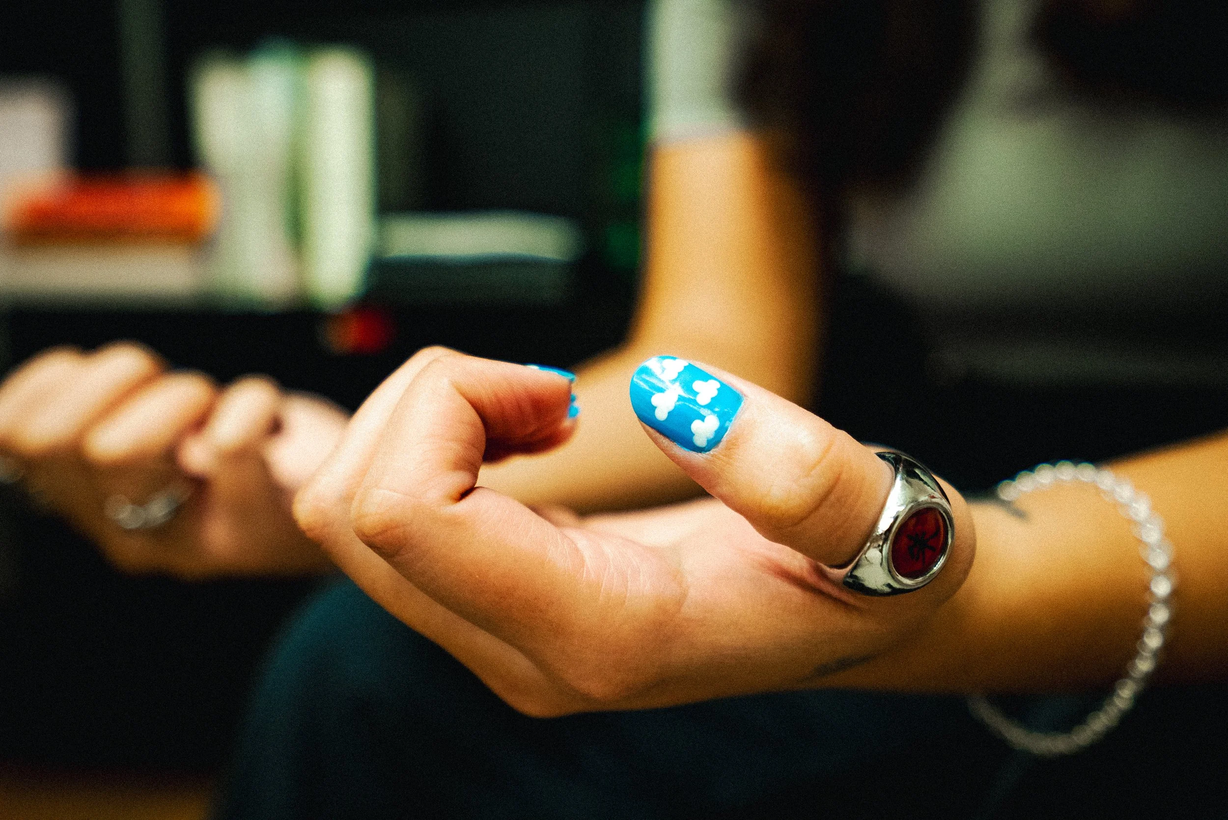 Close-up of a person's hand with blue nail polish and white floral designs, wearing rings, holding a small object with a blurred background.