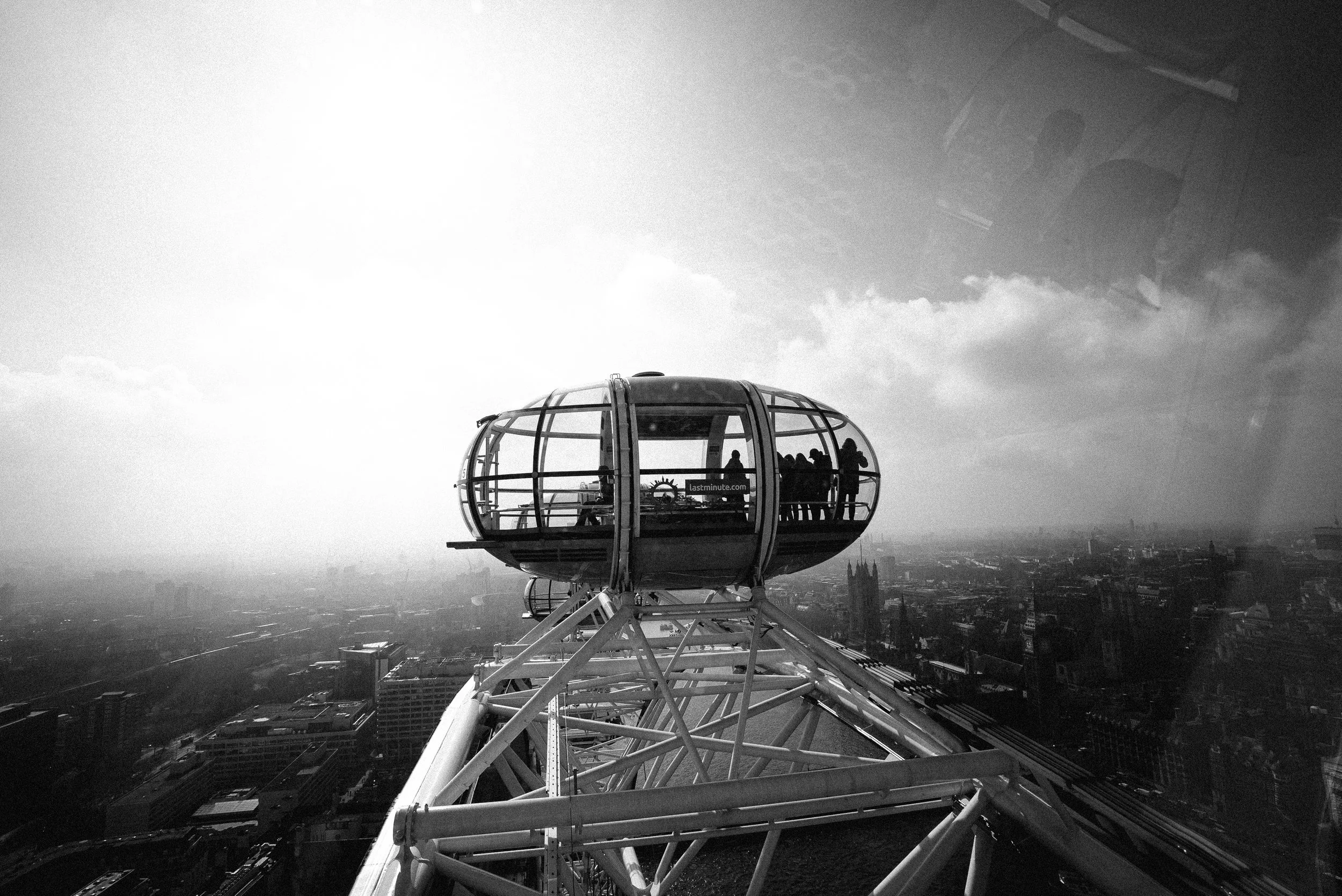 View of a Ferris wheel with glass-enclosed cabins, looking over a city skyline with tall buildings and landmarks in the background, under a cloudy sky.