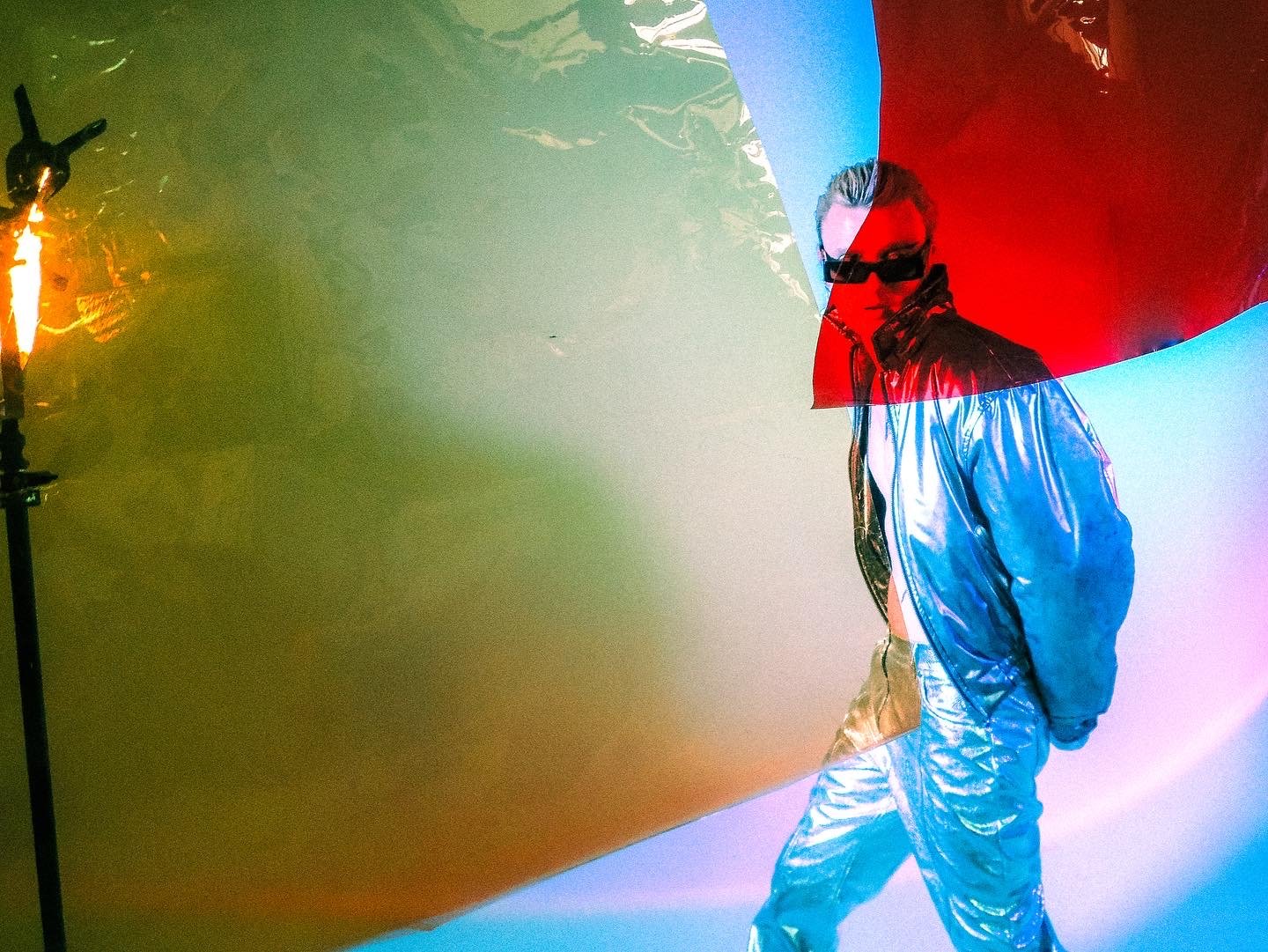 A person wearing reflective clothing and sunglasses stands in front of colorful, overlapping reflective surfaces, creating a layered, artistic effect.