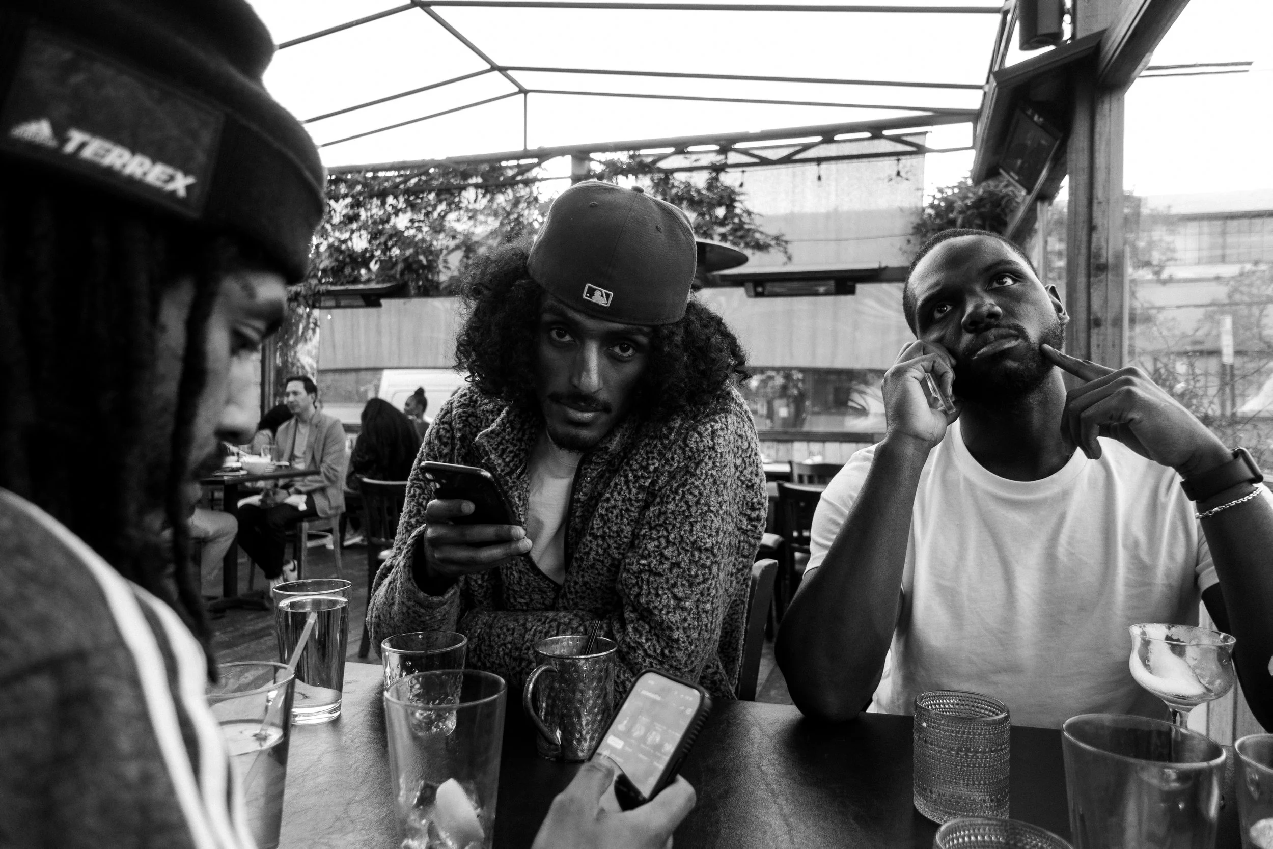 Black and white photo of three people sitting at a food court table, with glasses and cups in front of them.  the center is wearing a hat and patterned sweater, looking directly at the camera while holding a phone. The man to her right is on the phon