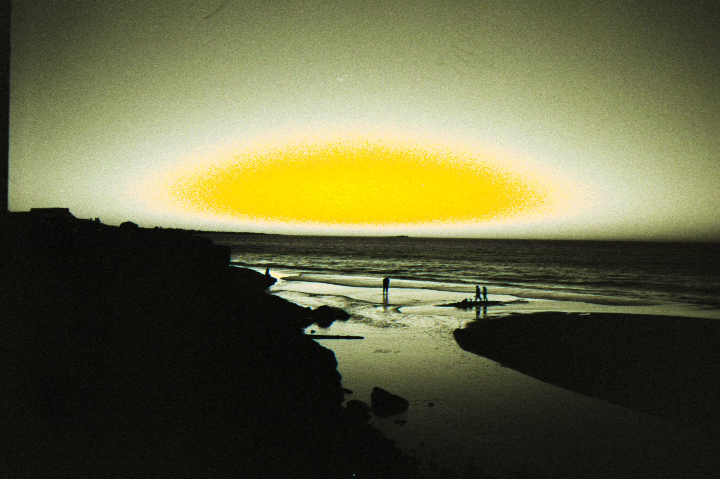 Sunset over the ocean with silhouettes of rocks, people walking on the beach, and a few birds in the sky.