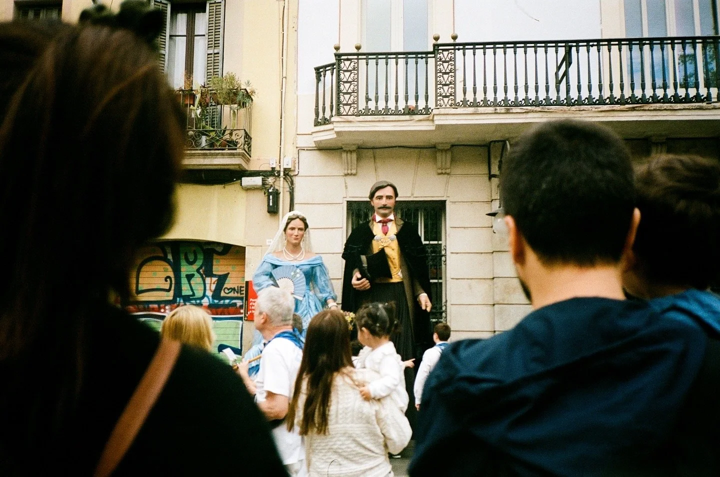 A street scene with a crowd watching a performance of actors dressed in historical costumes. The actors are on a raised platform in front of a building with balconies.