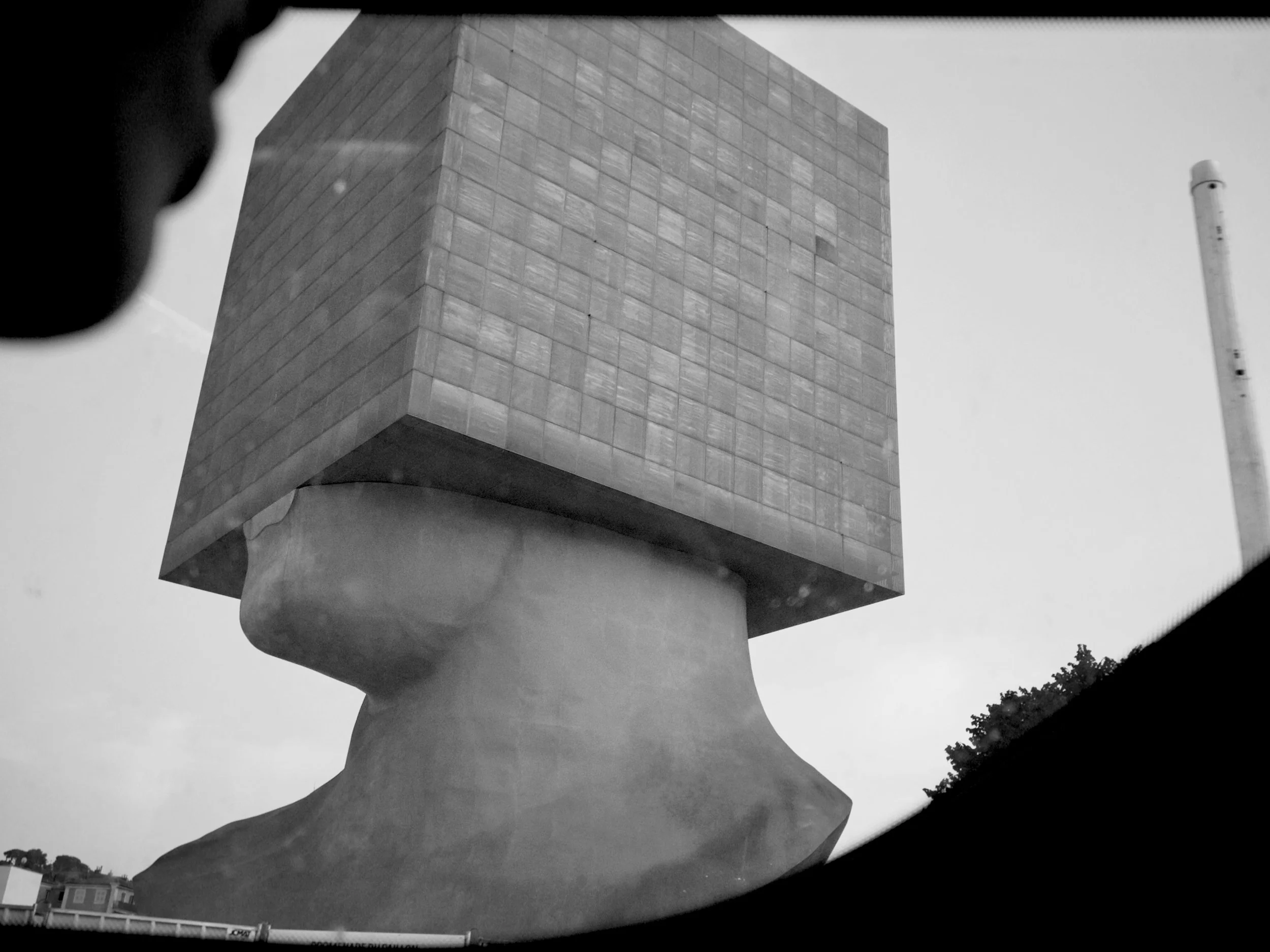 A black-and-white photo of a large modern sculpture of a human face profile, with a building structure on top, outdoors with trees in the background.