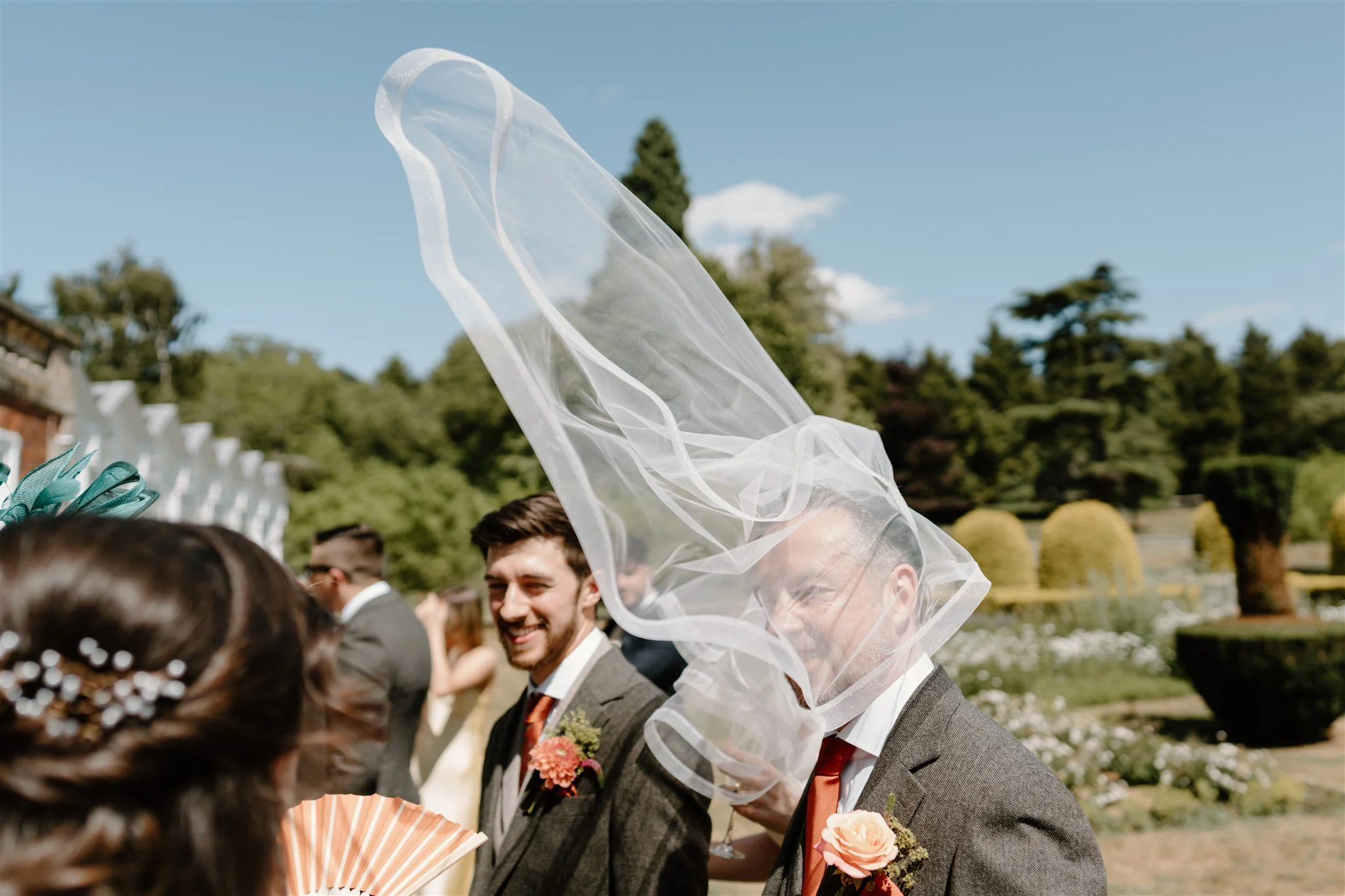 Documentary wedding photograph of a guest laughing as a long veil blows over his head during outdoor drinks