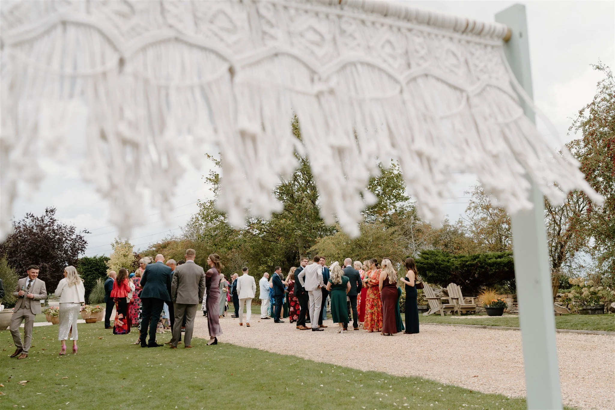 Candid wedding reception photograph of guests mingling in a garden, framed through a macrame backdrop