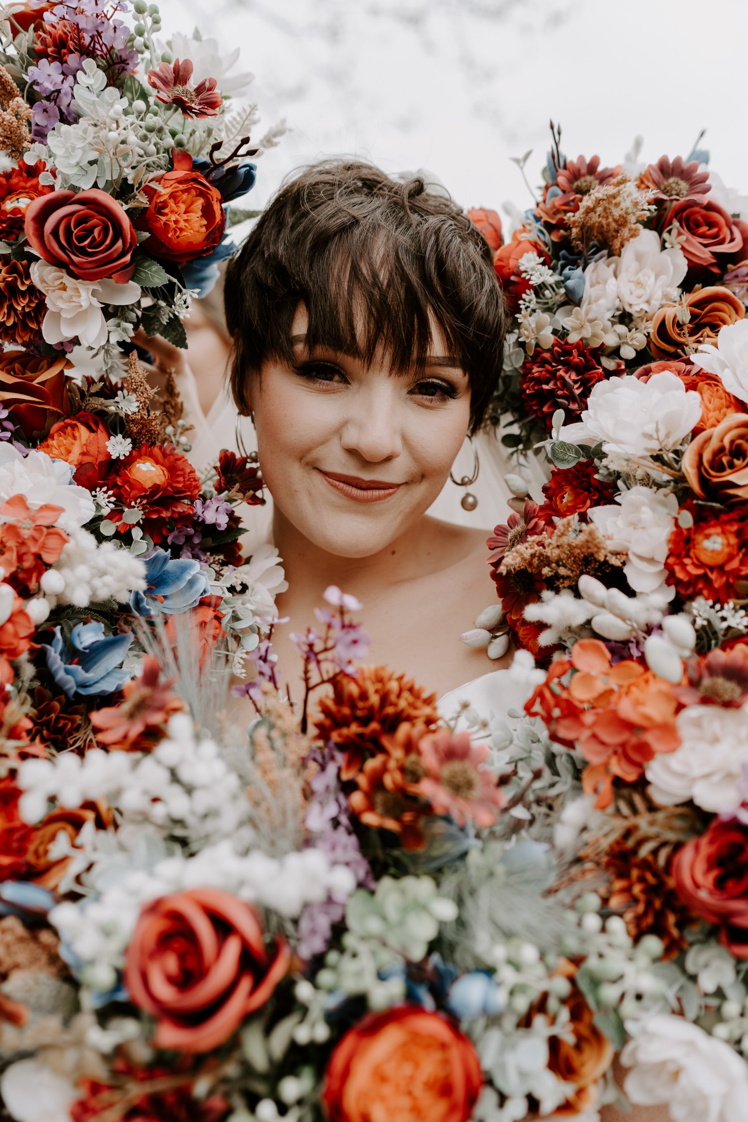 short haired bride surrounded by colourful orange red flower bouquets around head mardi gras village hall wedding Peterborough
