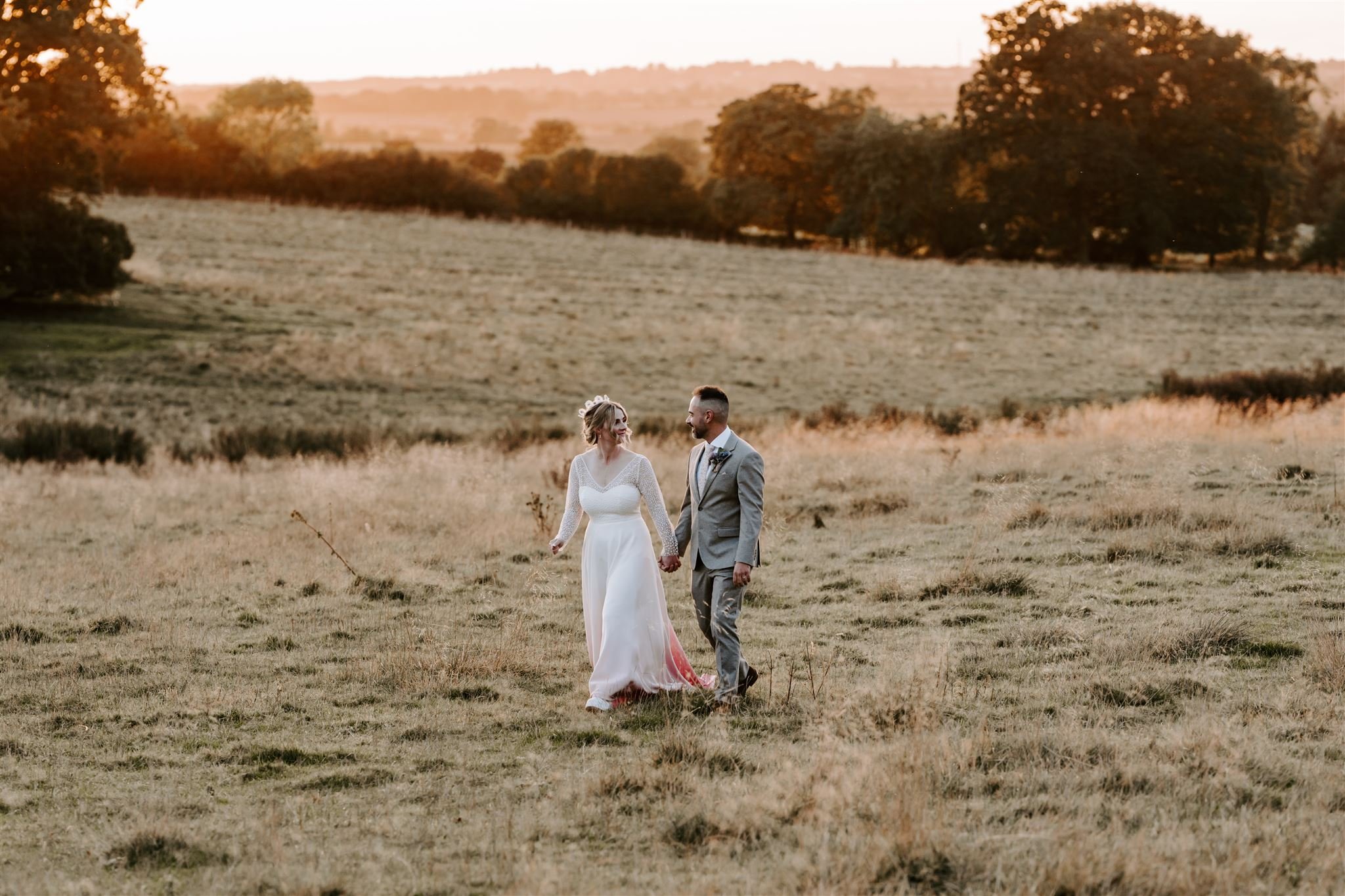 bride and groom walking in field on wedding day during a golden hour with bright orange skies and sun setting behind them Buckminister Barns wedding 