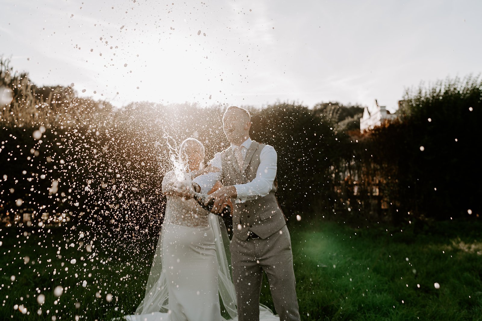 couple spraying champagne during golden hour with sun setting behind them and highlighting the champagne droplets 