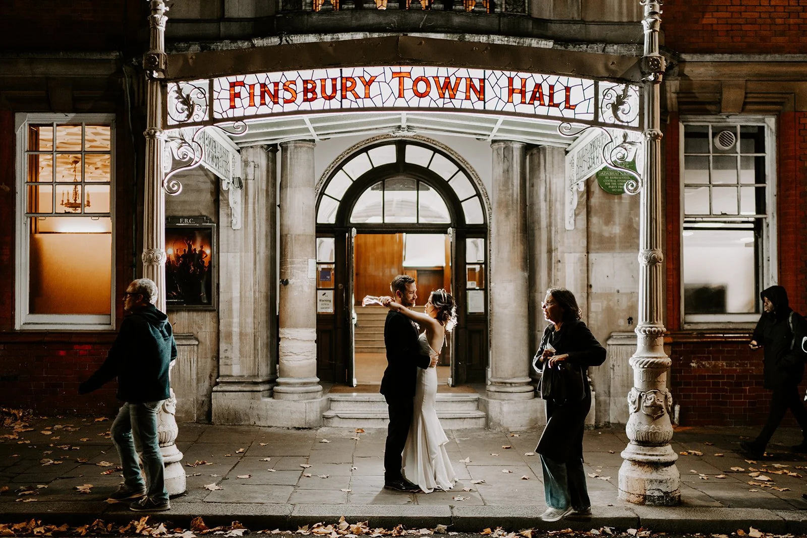 Couple standing outside Finsbury Town Hall at night while London traffic and pedestrians move around them