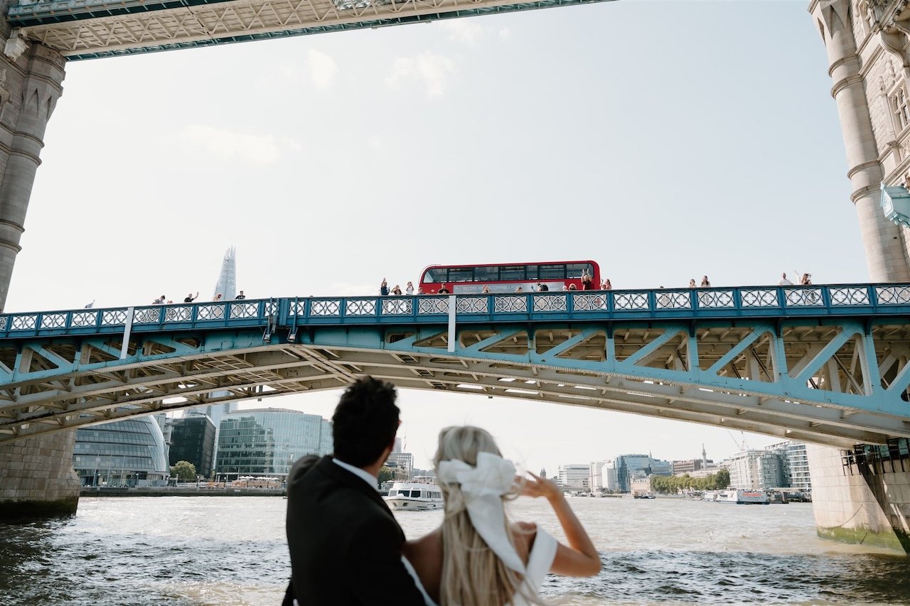 Bride and groom waving at guests from a boat passing under Tower Bridge