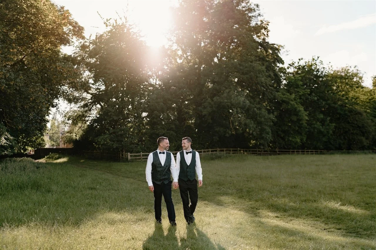 Two grooms walking together in the fields at The William Cecil in Stamford, relaxed documentary wedding photography at sunset