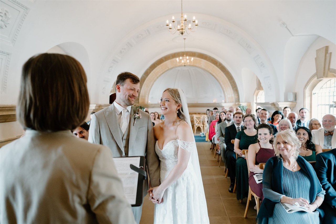 Wedding ceremony at Normanton Church in Rutland, with the couple smiling and holding hands, facing the celebrant and their guests.