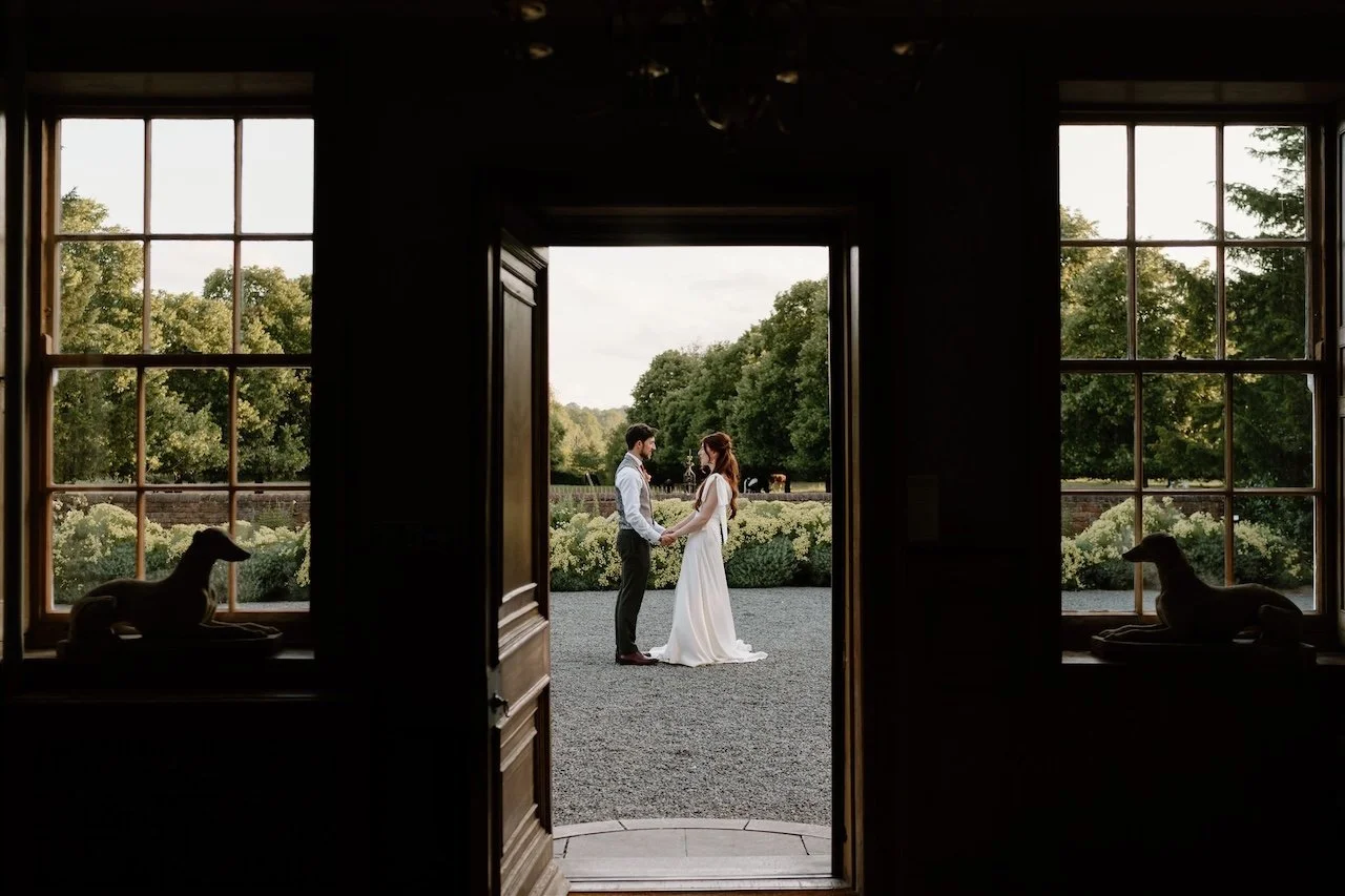 Bride and groom holding hands outside Sutton Bonington Hall, framed through open doors