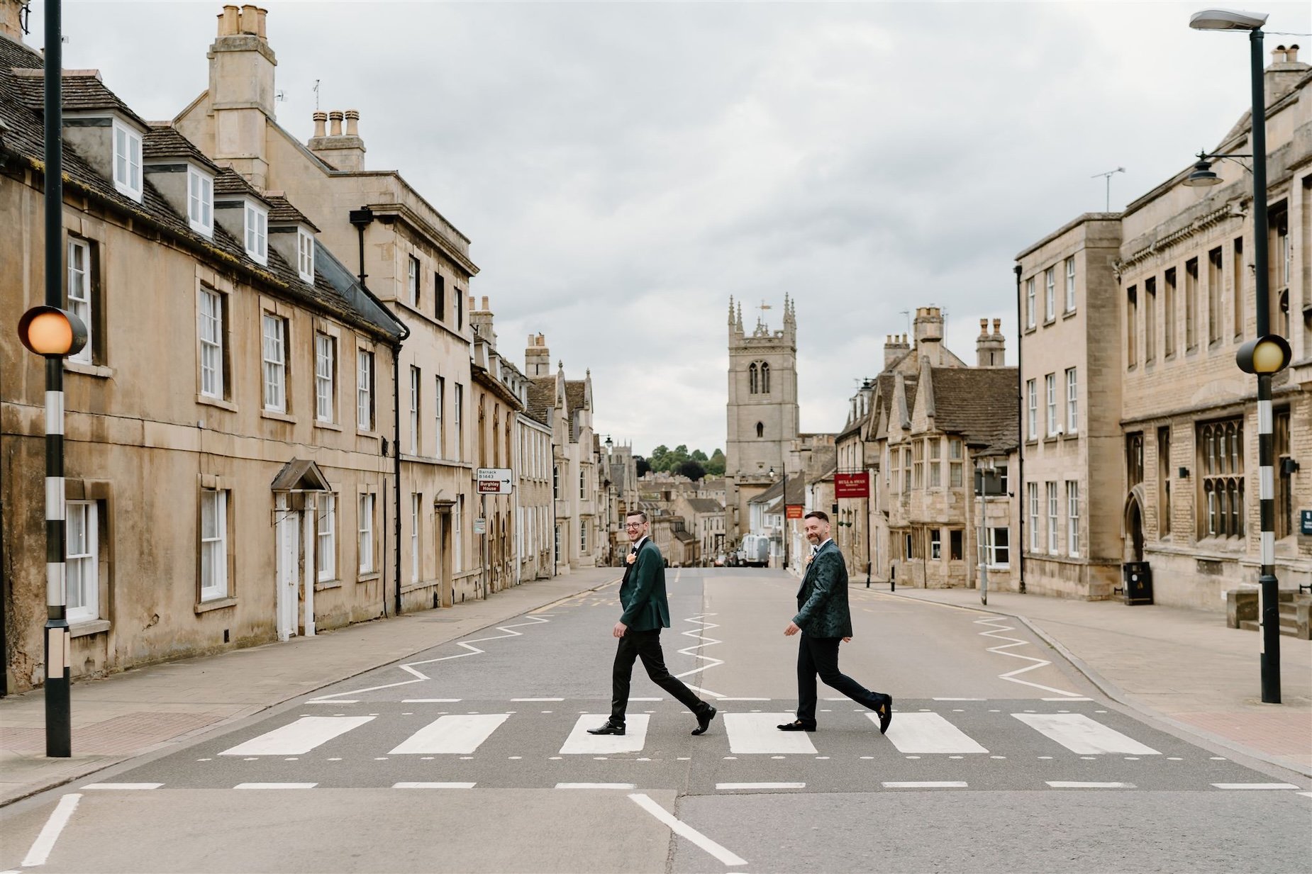 Two grooms walking across a pedestrian crossing in Stamford with historic stone buildings in the background.