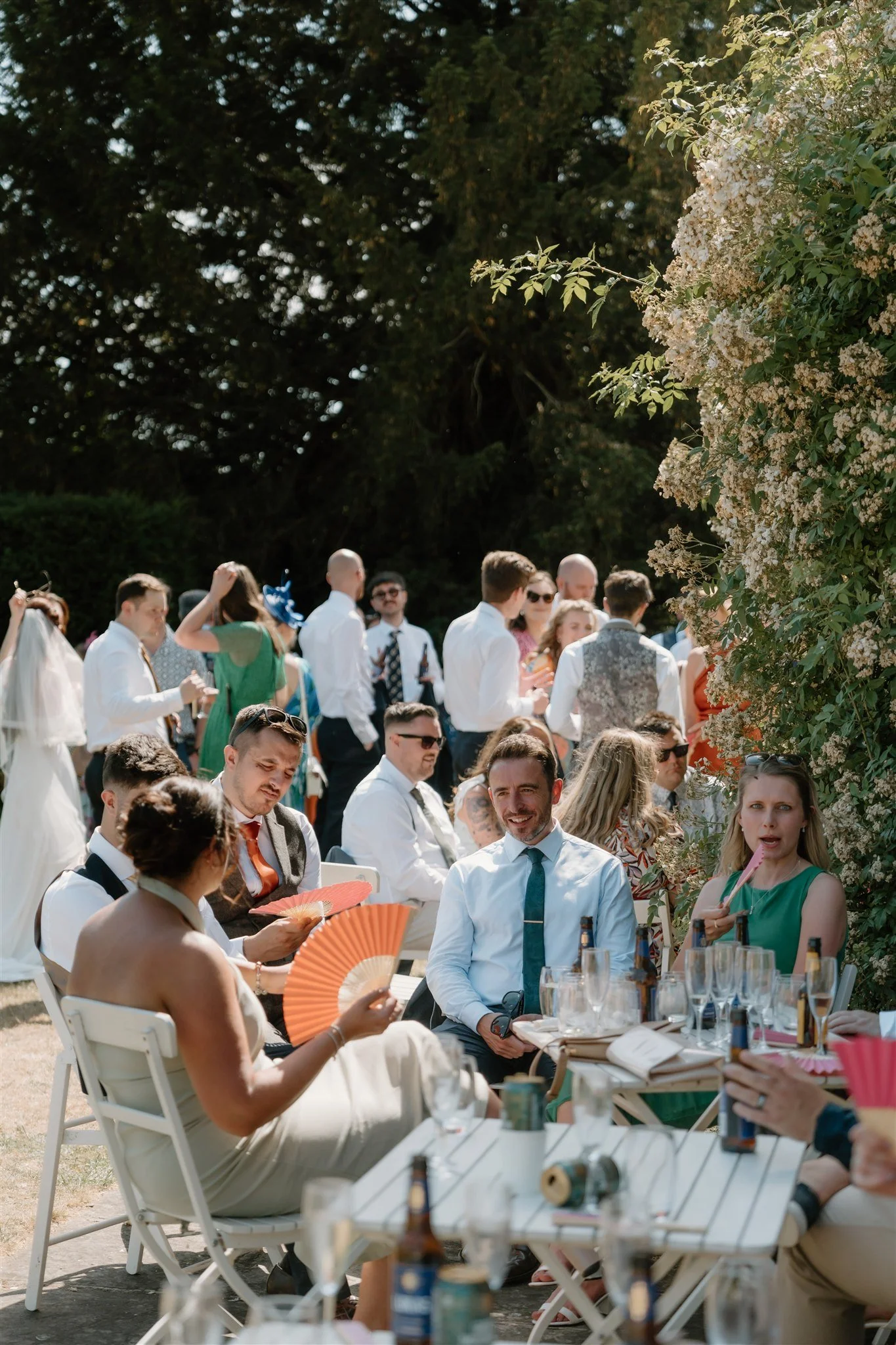 Candid photograph of guests talking and fanning themselves in the sun during an outdoor wedding reception, natural documentary wedding photography.