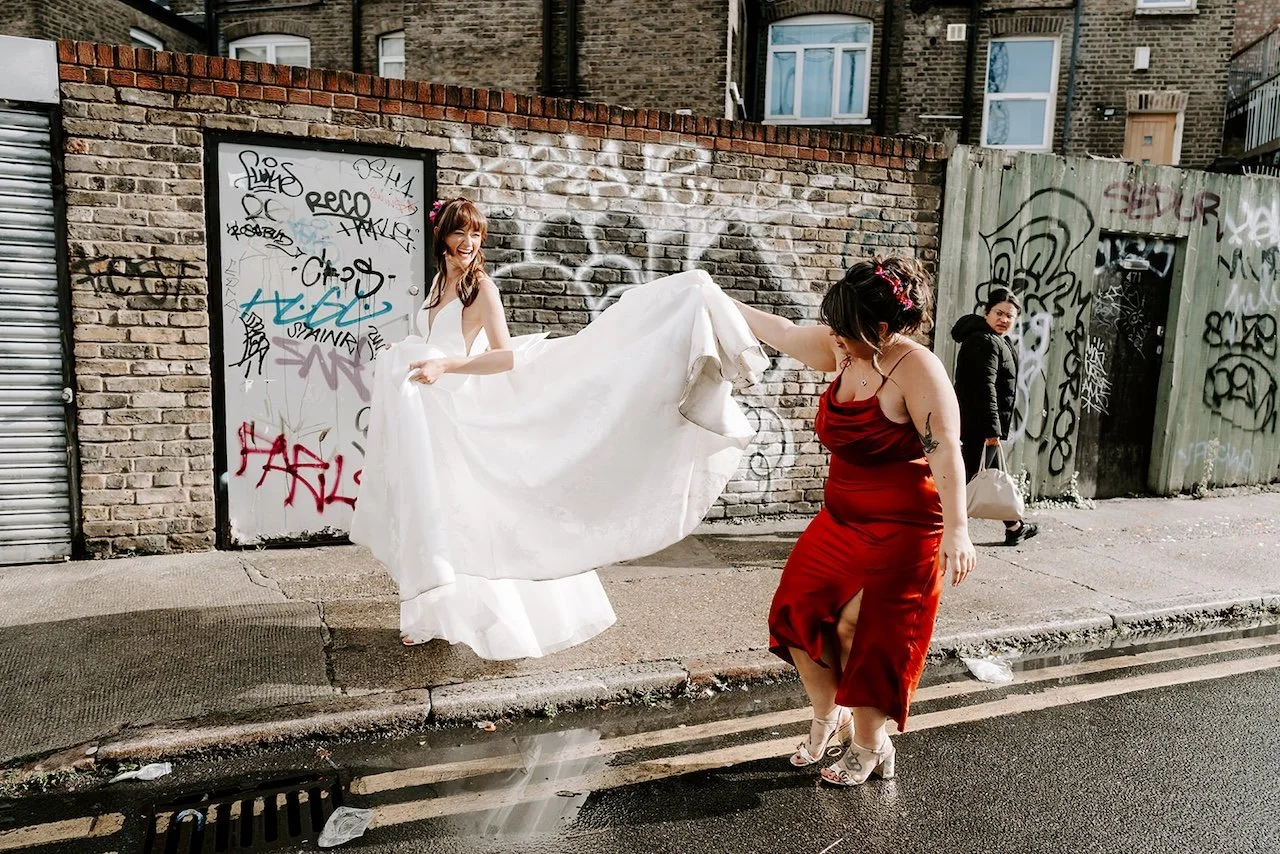 Bride laughing as a bridesmaid lifts her dress while walking along a graffiti covered street in Hackney, East London after the wedding.