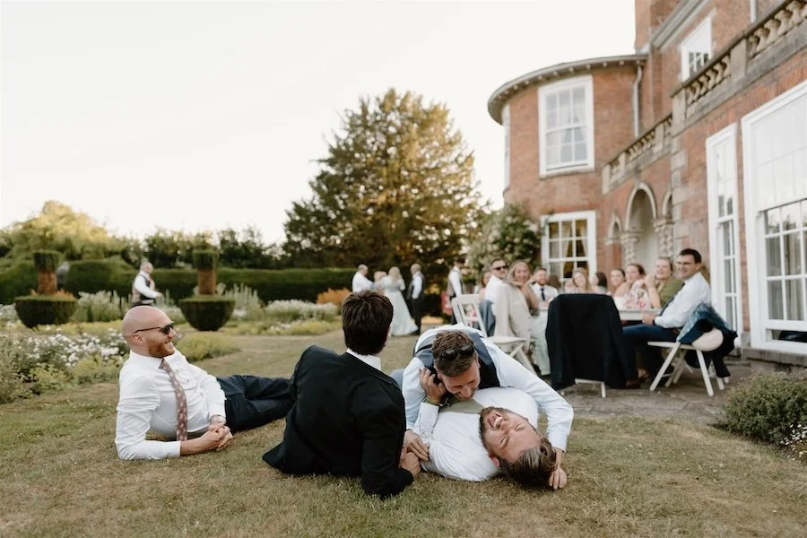 Candid wedding photograph of guests relaxing and play wrestling on the lawn of a country house reception