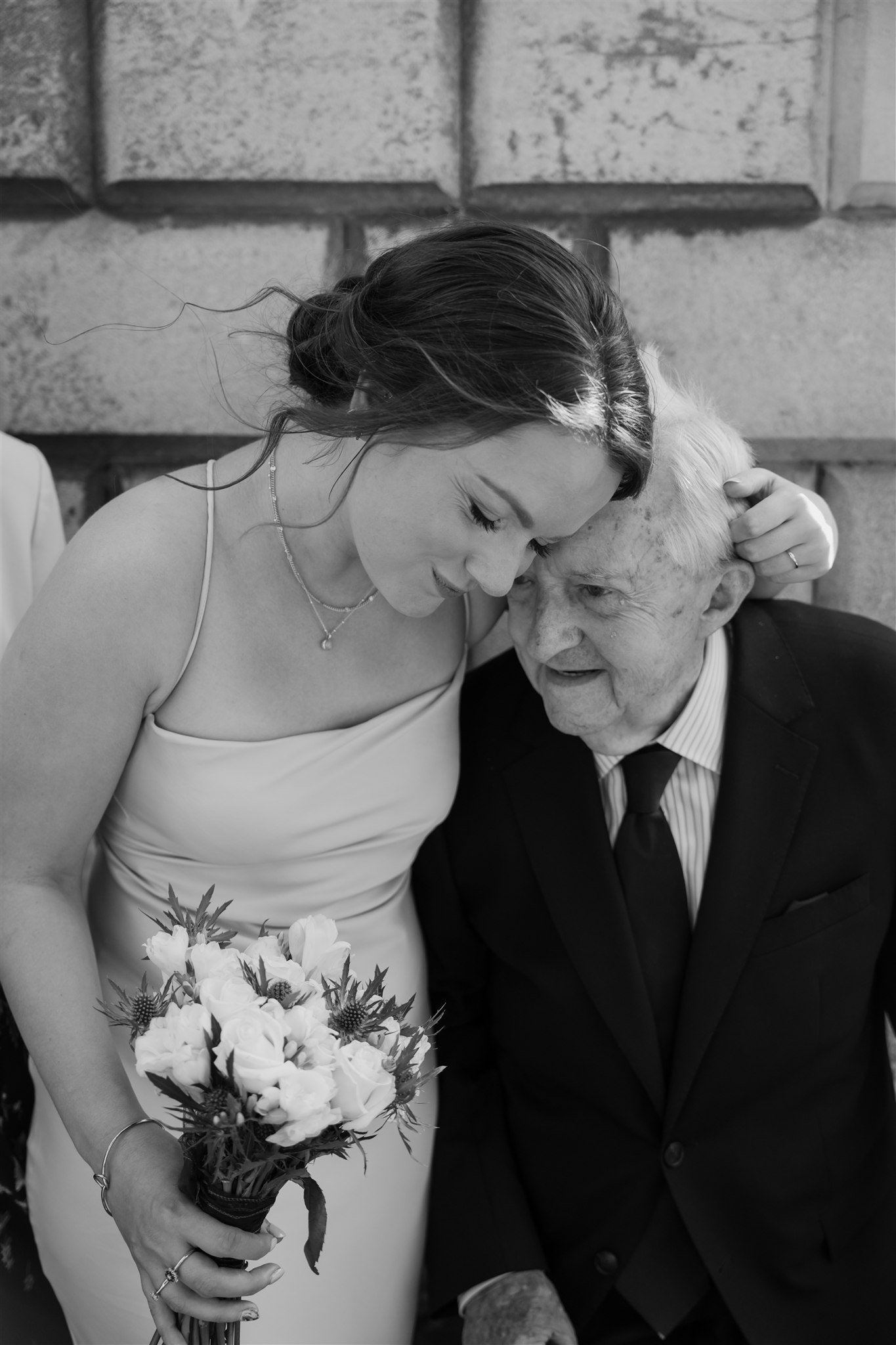 Black and white photograph of a bride resting her forehead against an elderly relative while holding her bouquet, intimate documentary wedding photography showing a generational moment.