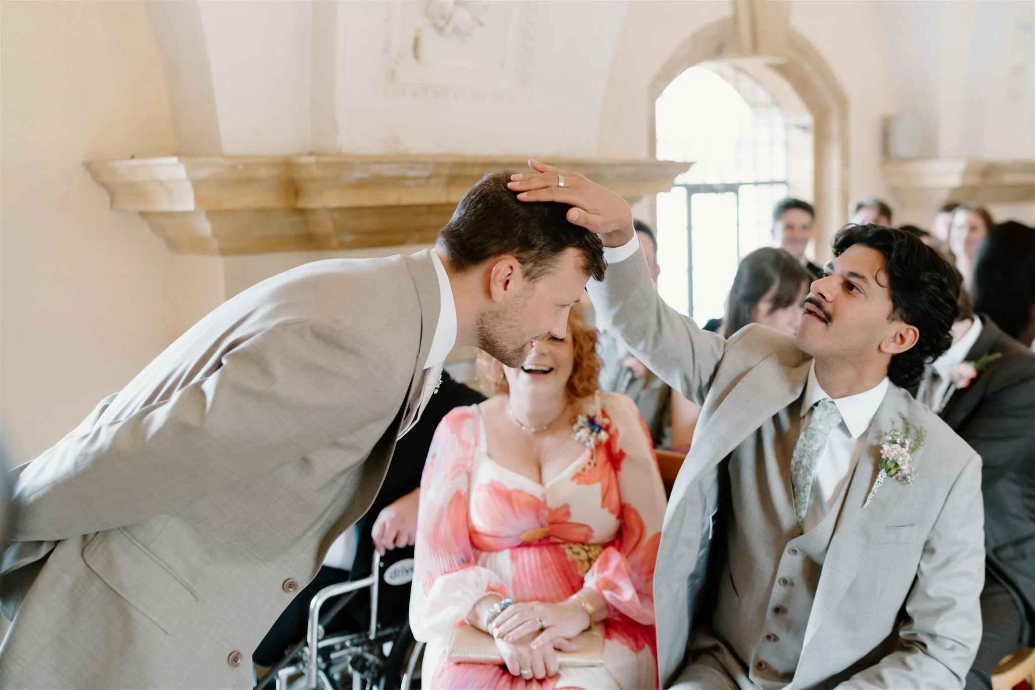 Wedding guest playfully patting groom's head during the ceremony while the woman between them laughs