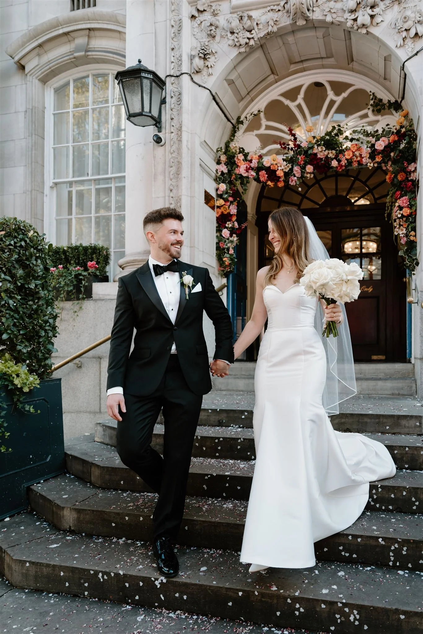 Couple walking down the steps of Chelsea Town Hall under a colourful flower arch, London town hall wedding