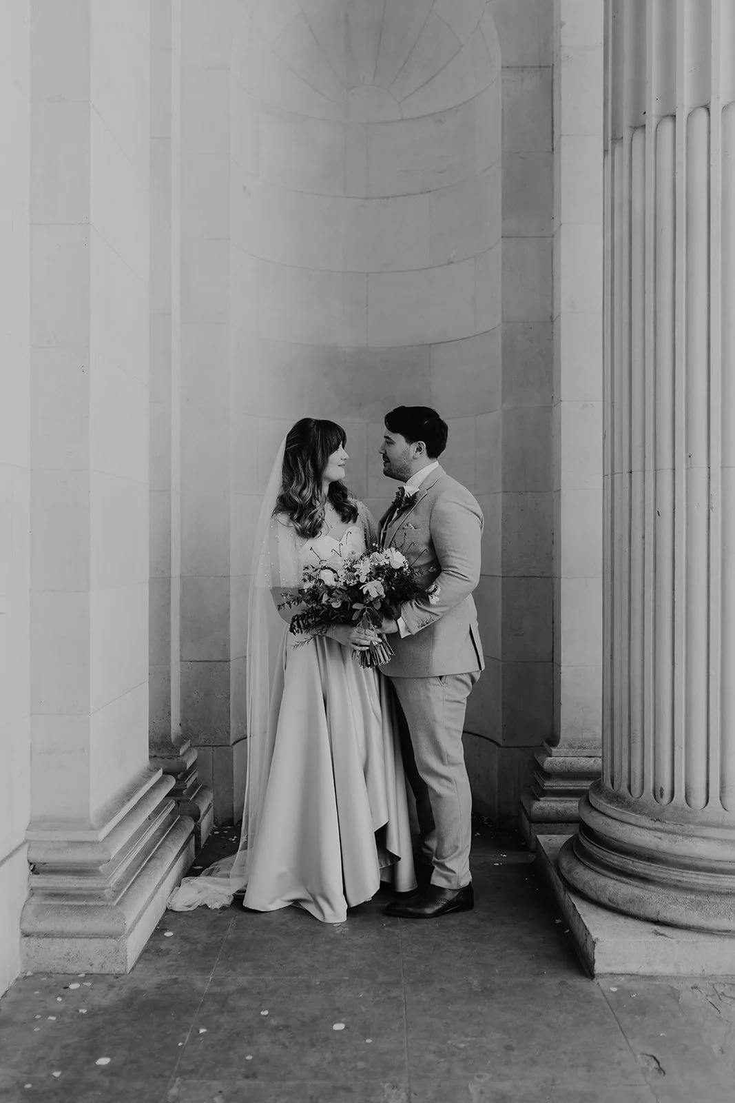 Bride and groom standing between the columns at Marylebone Town Hall in London, holding a bouquet and sharing a quiet moment after their ceremony