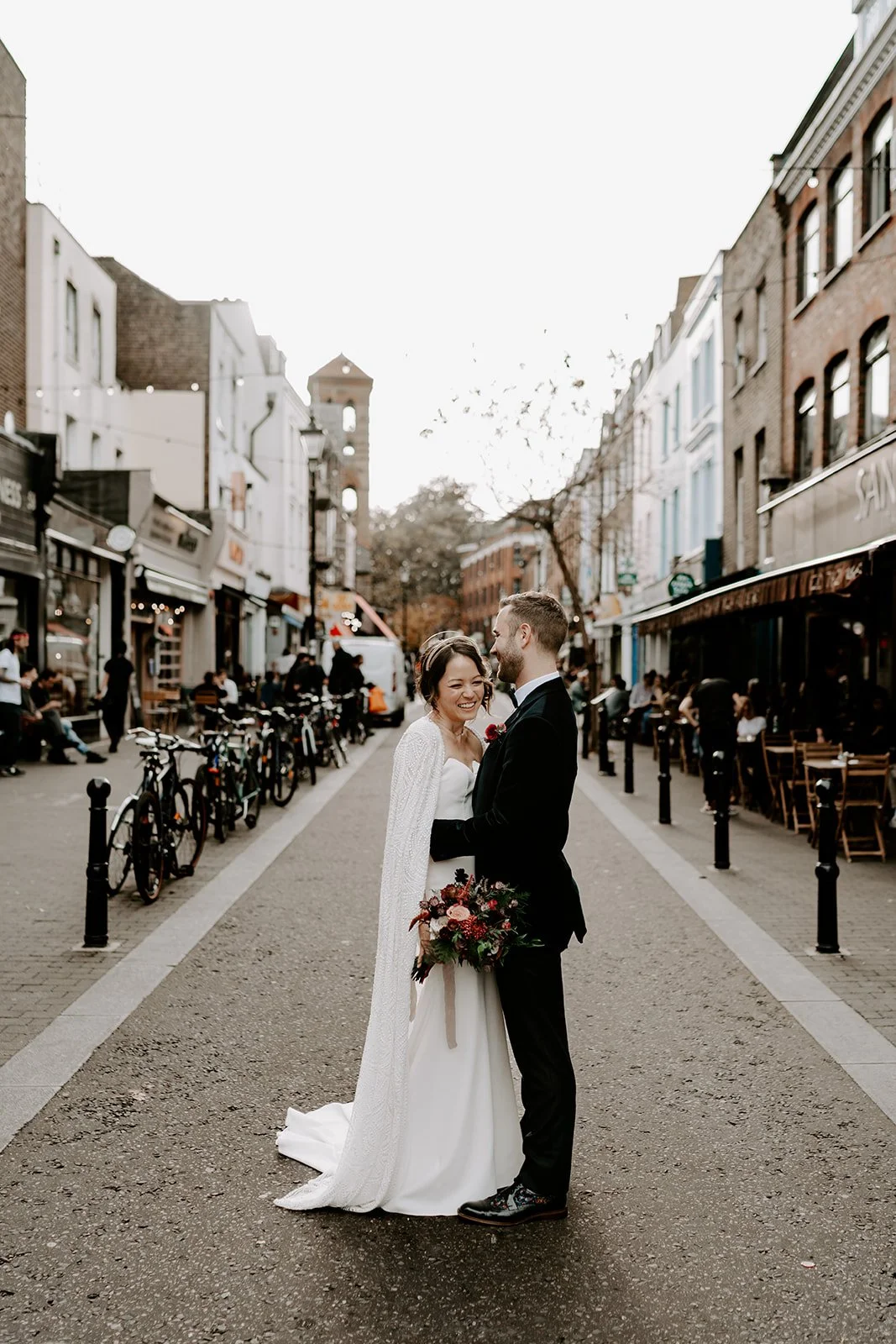 London wedding couple standing in the middle of a narrow street with cafés and bicycles on both sides