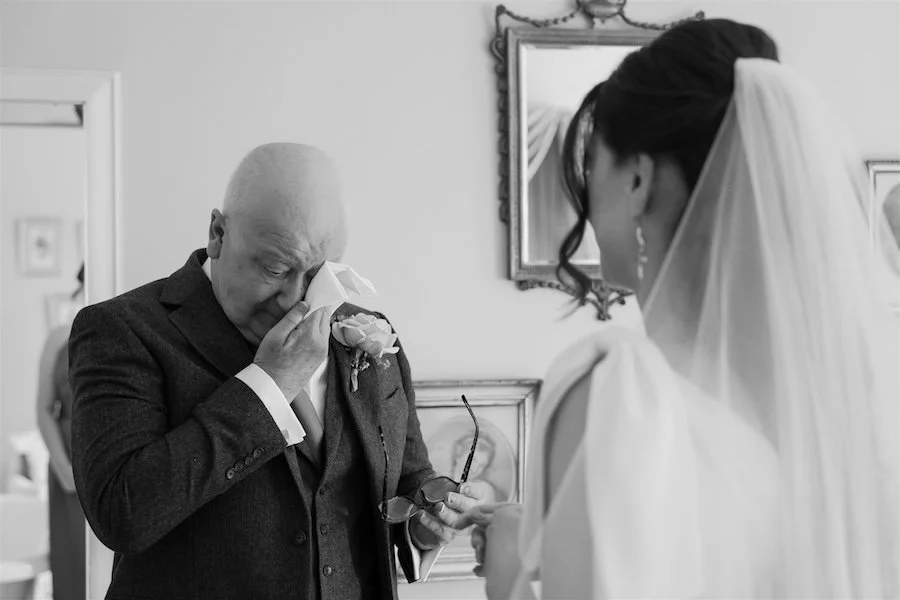 Black and white documentary wedding photograph of a father wiping away tears as he sees his daughter in her dress