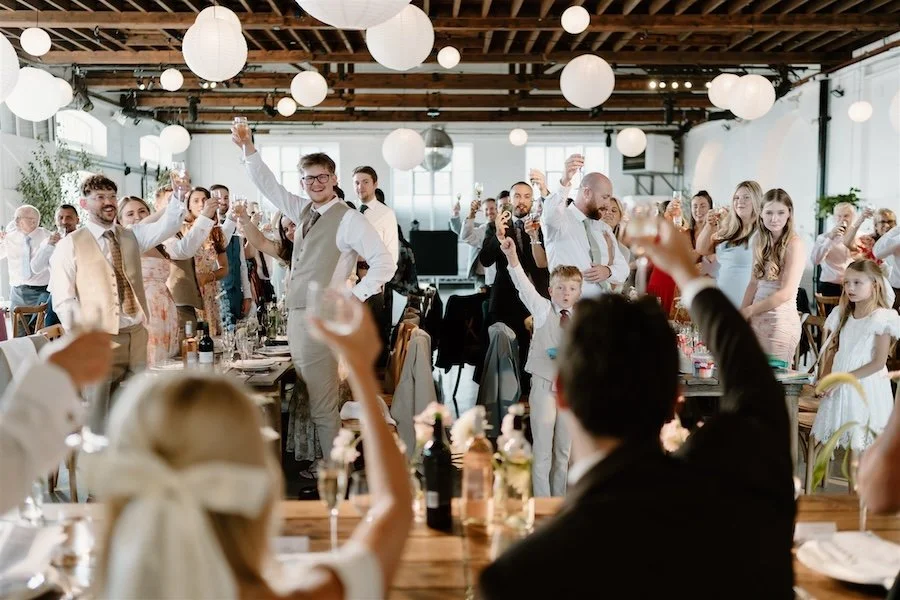 Guests stand and raise their glasses during wedding speeches, photographed from the couple’s table
