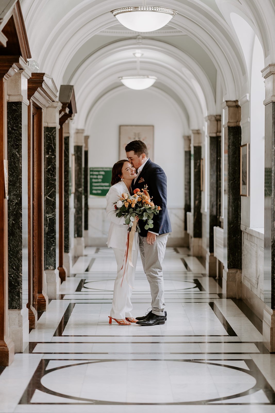 Bride in a white suit with her partner standing in the marble corridor at Islington Town Hall, London wedding portraits