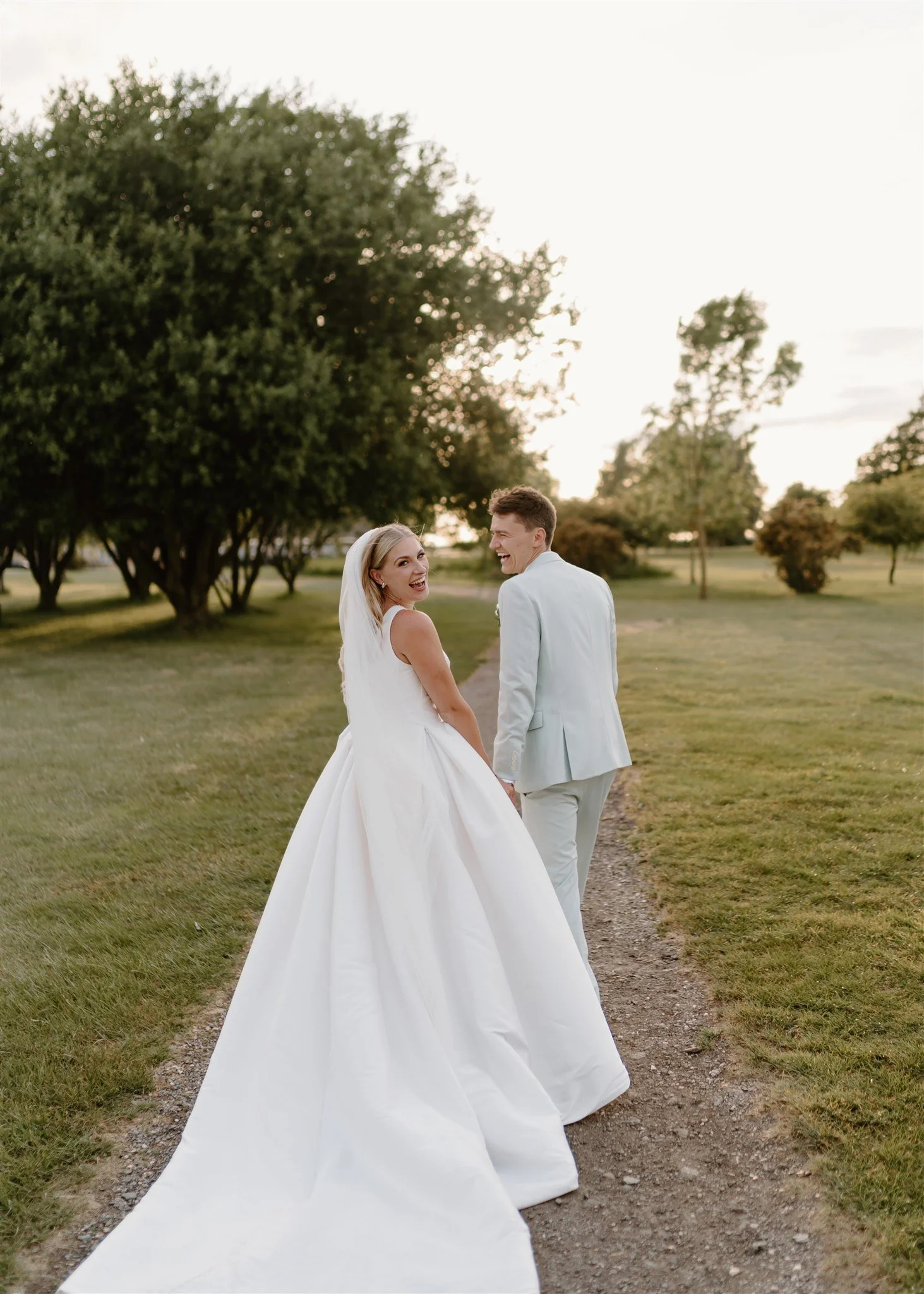 Bride in a full skirted gown walking hand in hand with her groom along a path at sunset, relaxed editorial style wedding photography.