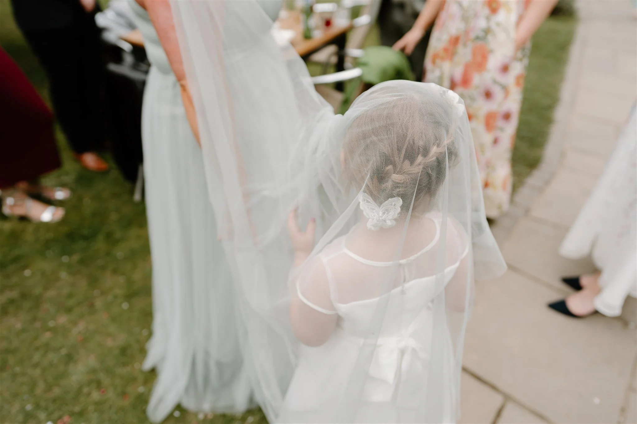 Flower girl standing under a long veil during wedding reception, photographed from behind in a soft documentary style