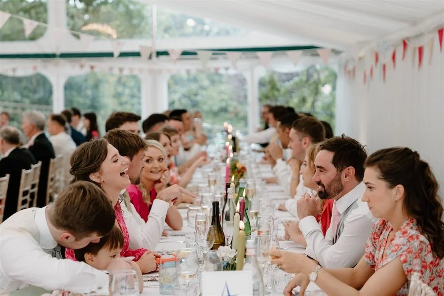 Documentary wedding reception photograph of guests laughing along a long marquee dinner table