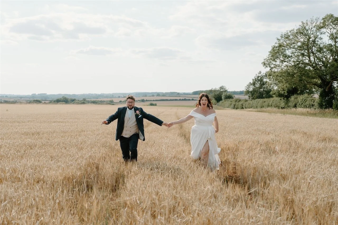 Bride and groom running hand in hand through a golden field near Ed’s Hill Top marquee in Rutland and Stamford during their festival-style wedding