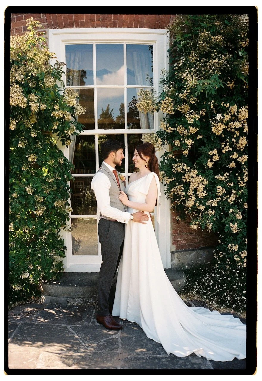 Bride and groom embracing in front of a window covered in climbing flowers at Sutton Bonington Hall.