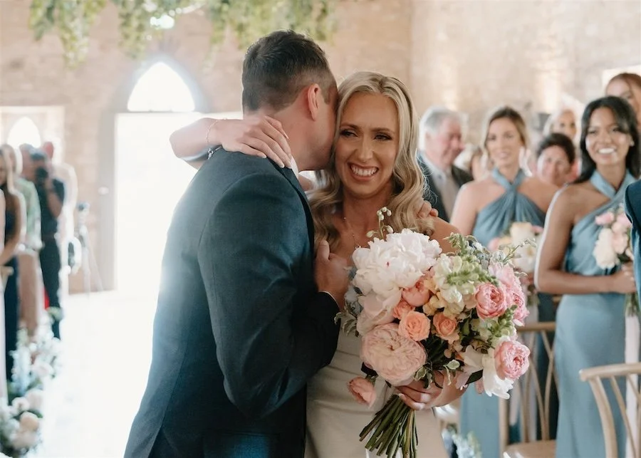 Bride smiles as she is hugged during the ceremony, holding a bouquet of pastel flowers with guests watching behind