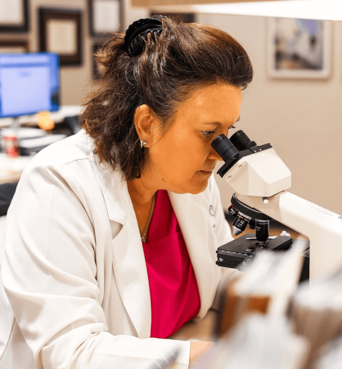 A woman in a white lab coat looks through a microscope in a laboratory setting.