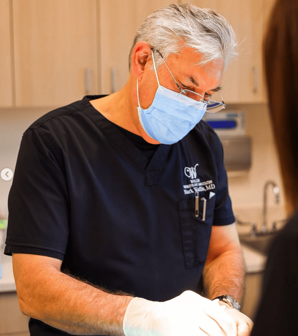 A male healthcare professional with gray hair, wearing a black medical scrub shirt with a logo, a face mask, and glasses, working in a clinical setting.