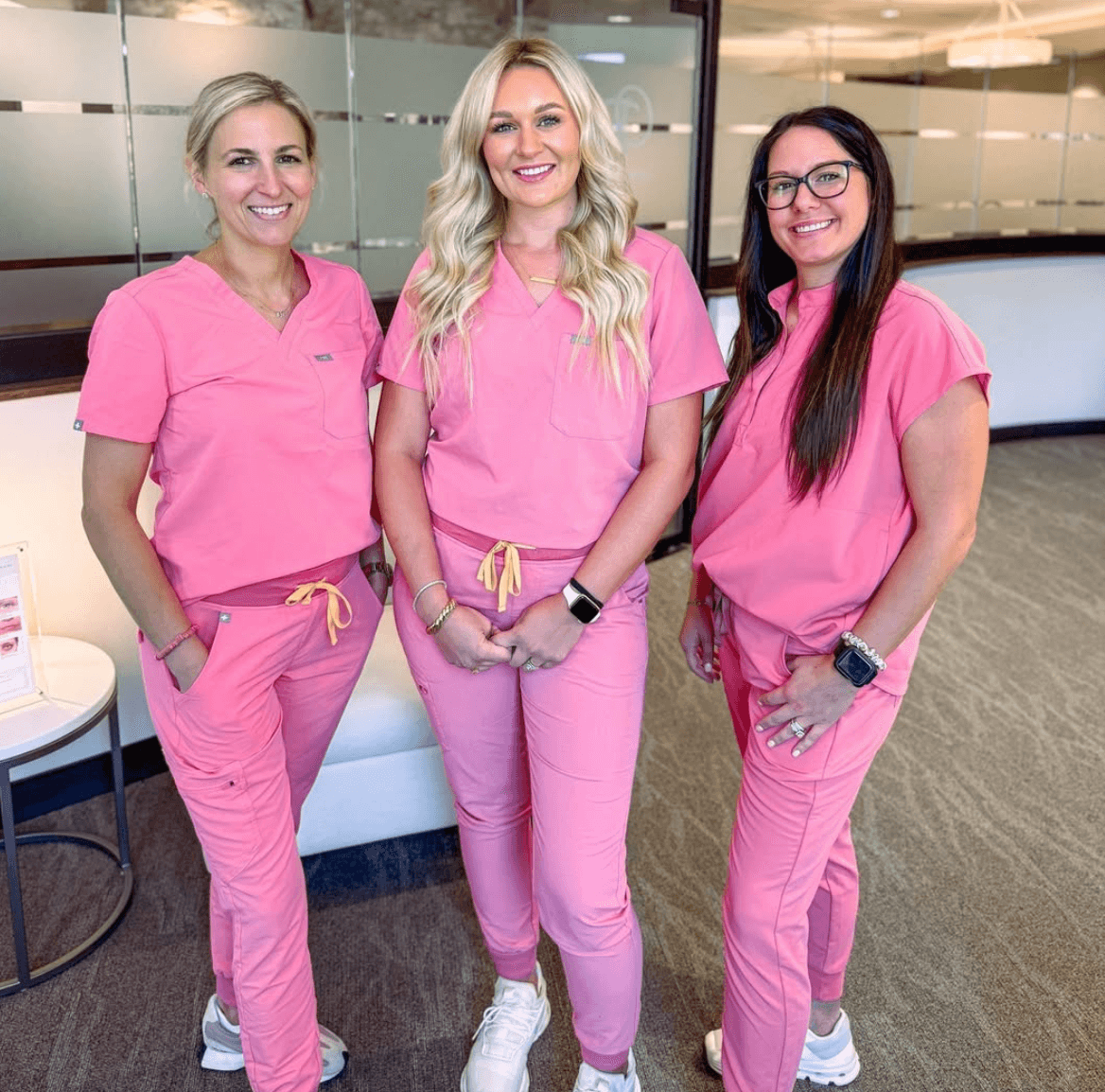 Three dental professionals in pink scrubs standing in an office, smiling at the camera.