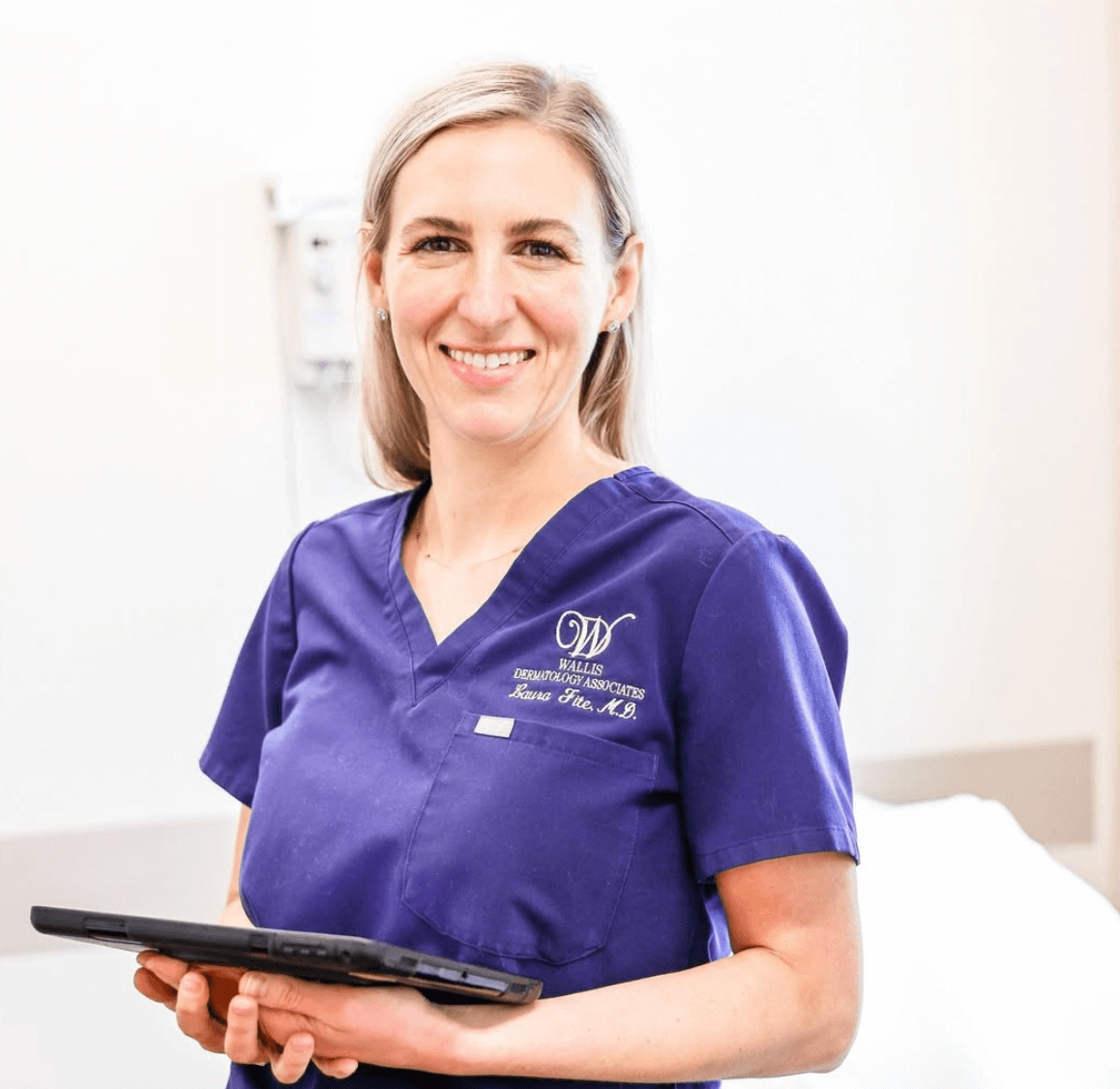 A smiling woman in purple medical scrubs holding a tablet, standing in a clinical setting.
