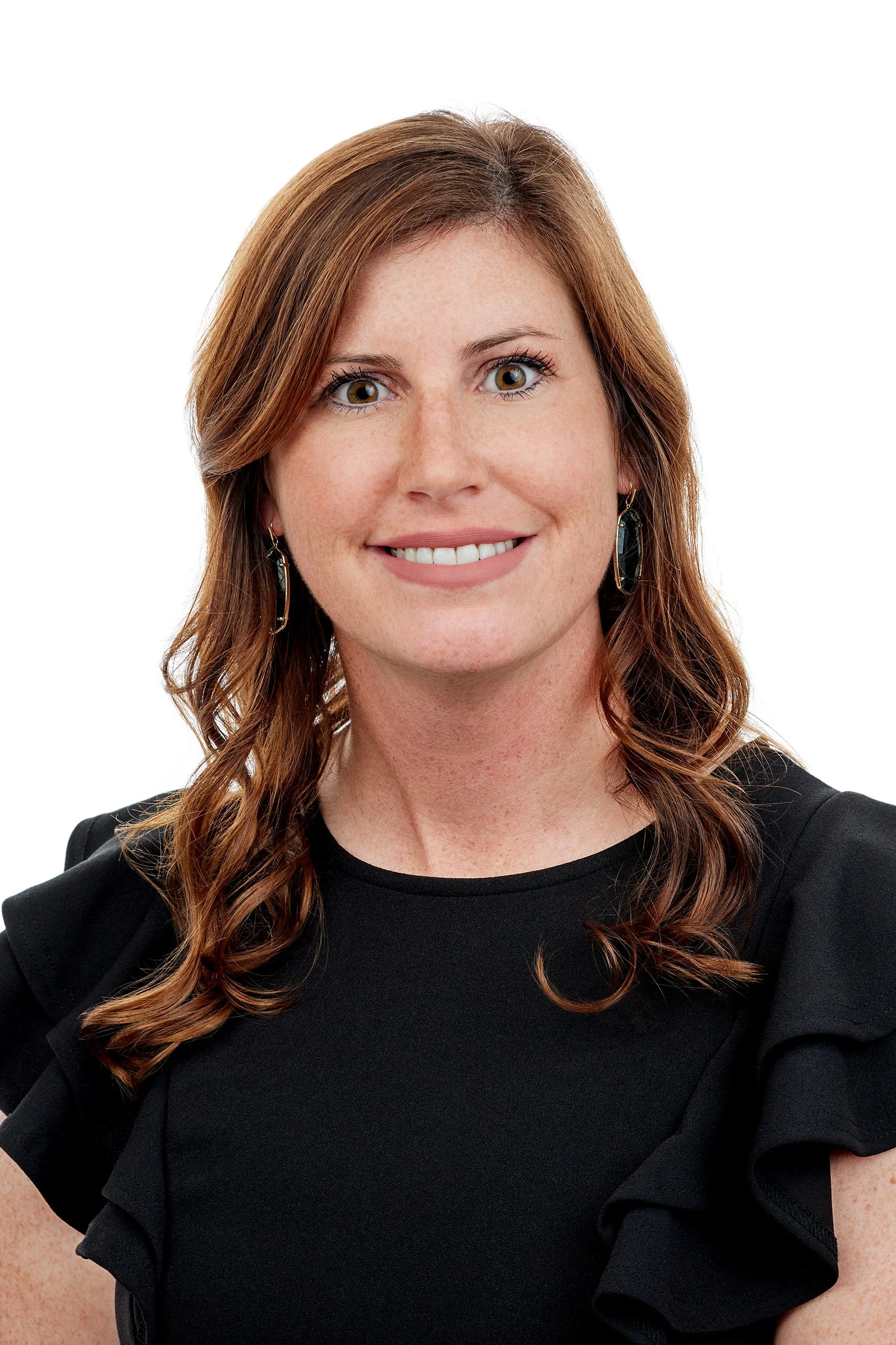 A woman with light skin, shoulder-length wavy brown hair, and green eyes, smiling, wearing a black top with ruffled shoulders and gold and black earrings, against a white background.