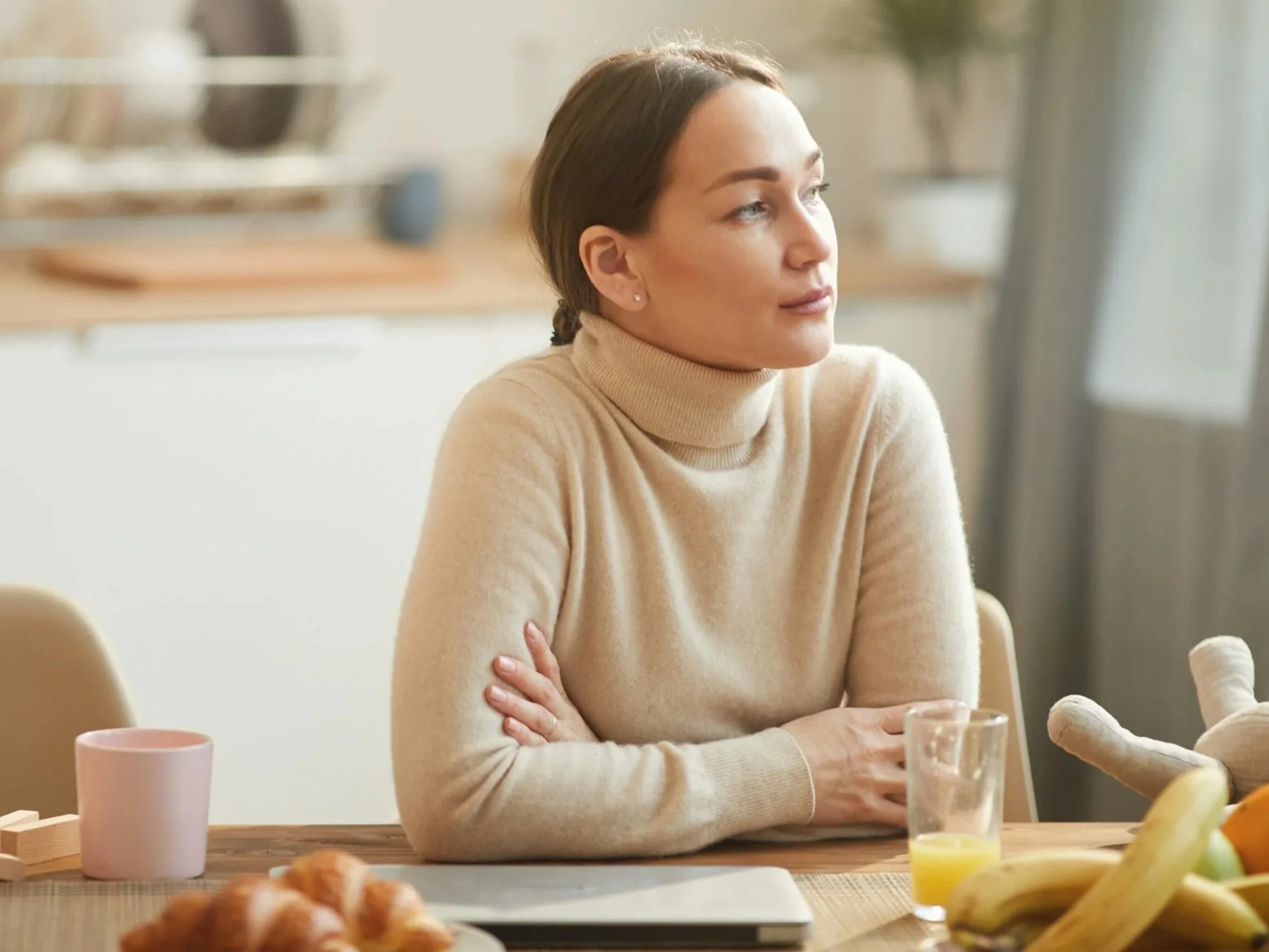 A woman sits at her table with a glass of orange juice, fruit, and croissants in front of her. She looks pensively off to the side.