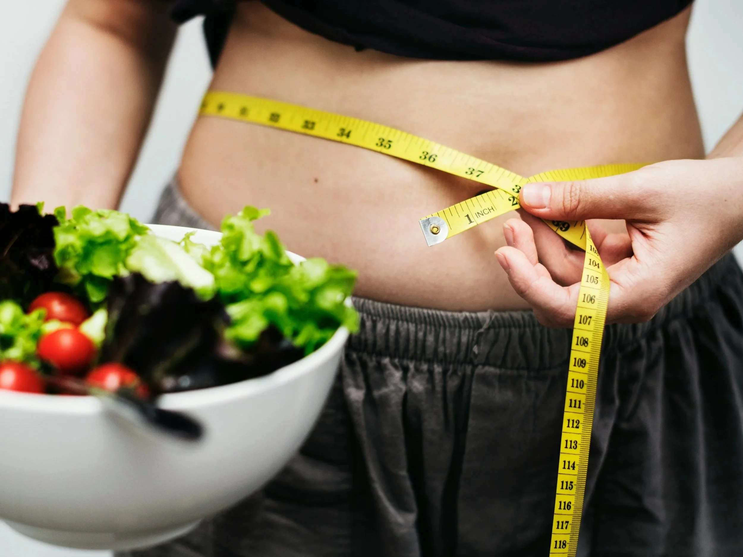 A close up of a woman's torso with her shirt pulled up and a measuring tape around her waist. One hand hods the measuring tape closed on 38 inches while the other hand holds a bowl full of salad, lettuce, and tomatoes.