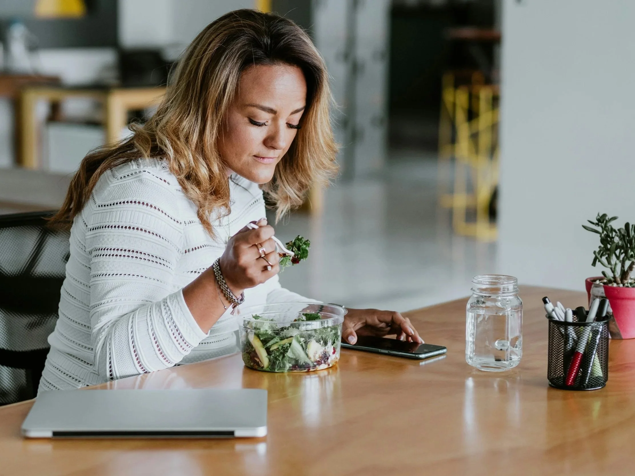 a woman sits at her desk with her laptop closed eating a fresh salad with a glass of water.