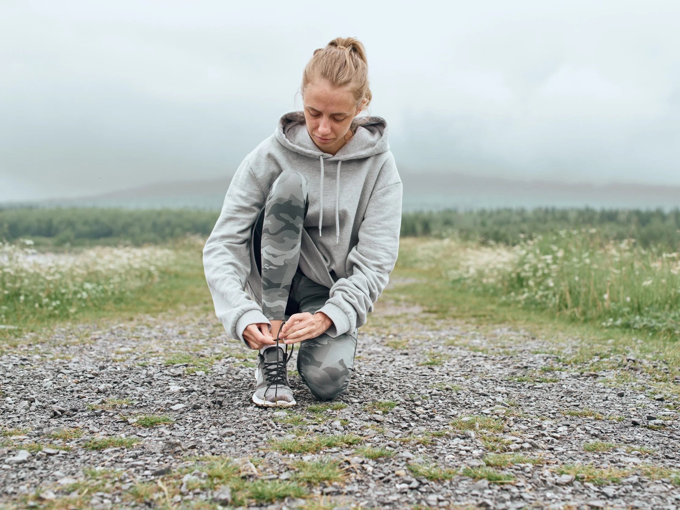 A woman looks pensive while lacing up her shoes in a field.